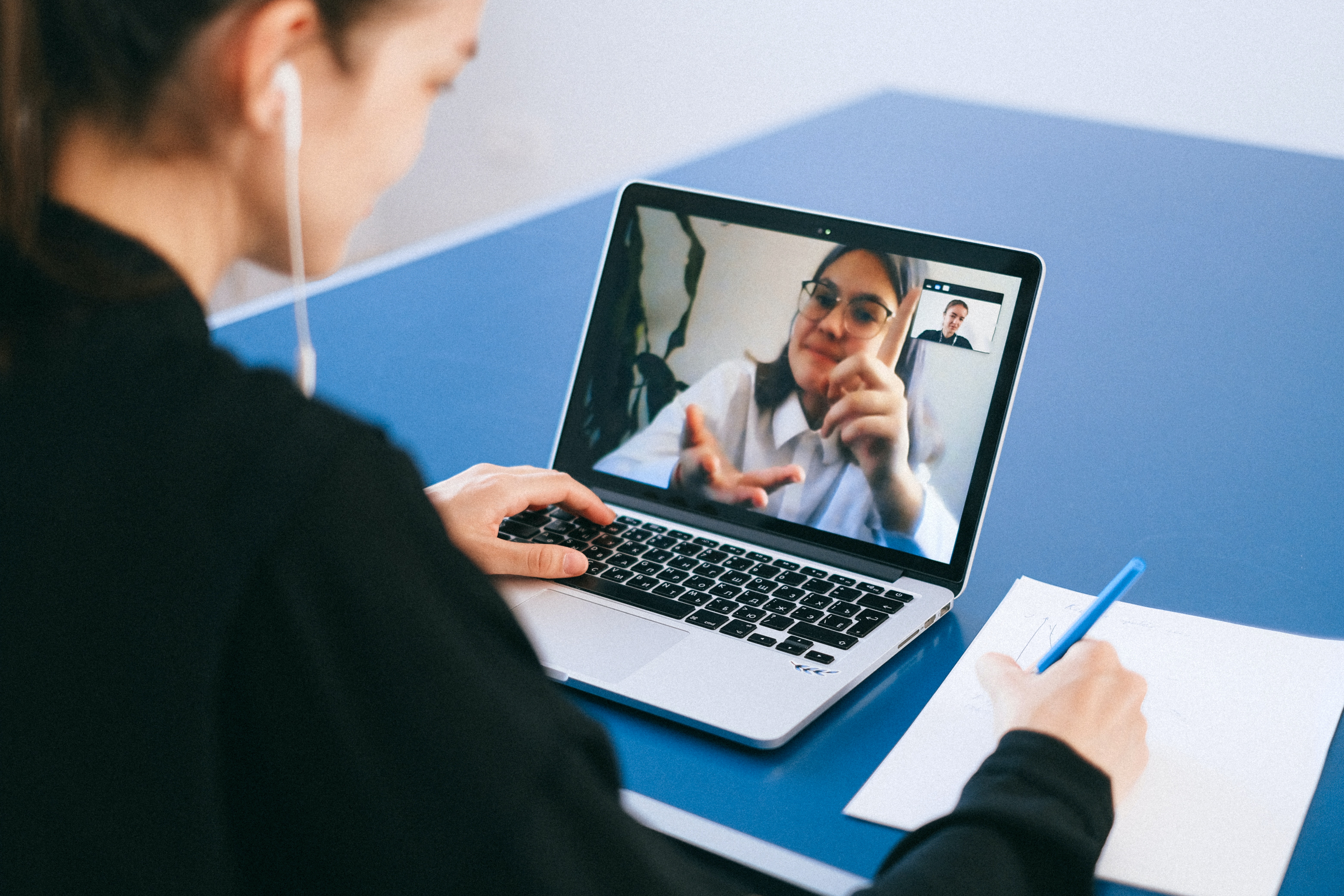 A woman in a black jacket sat at a table in front of a laptop making notes. Another woman in glasses is visible on the laptop screen as part of a video call.