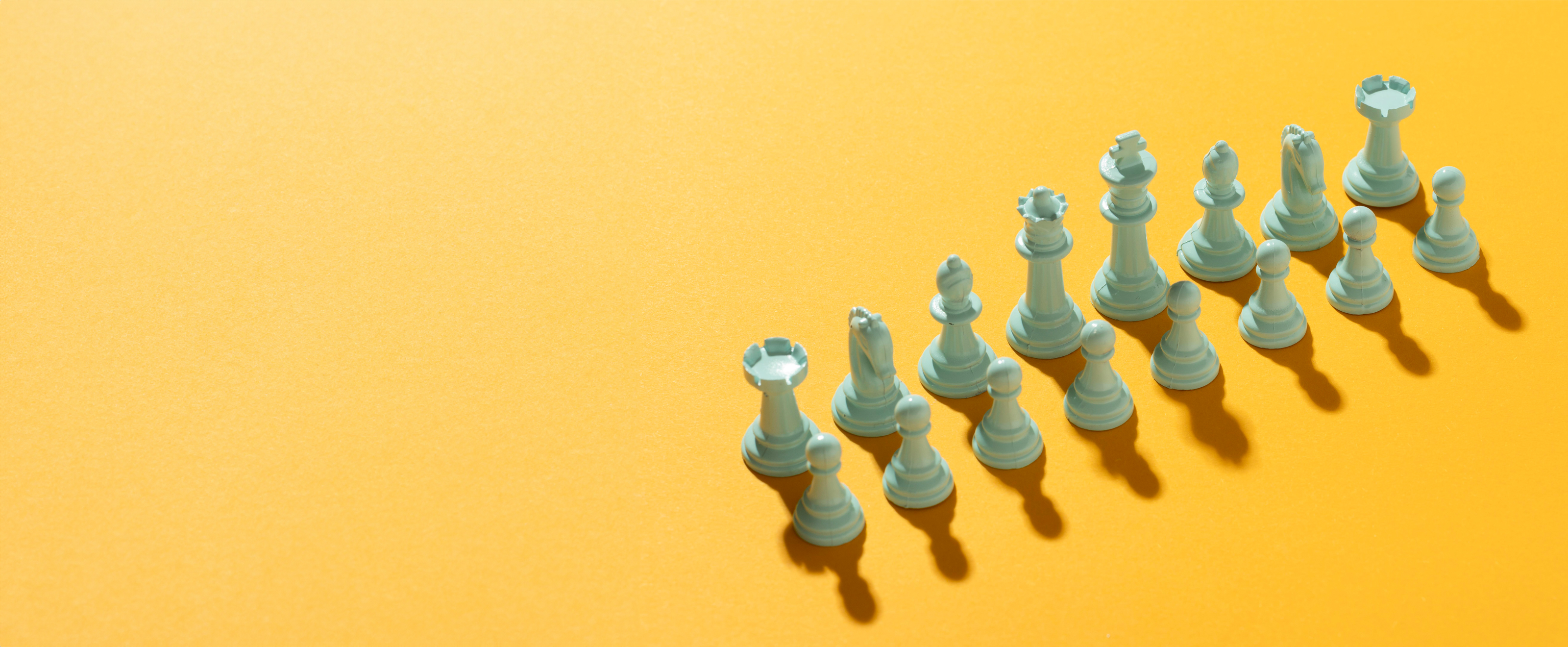 Photograph of a full set of white chess pieces, arranged as they would be for a new game, on a yellow background with strong shadows for each piece