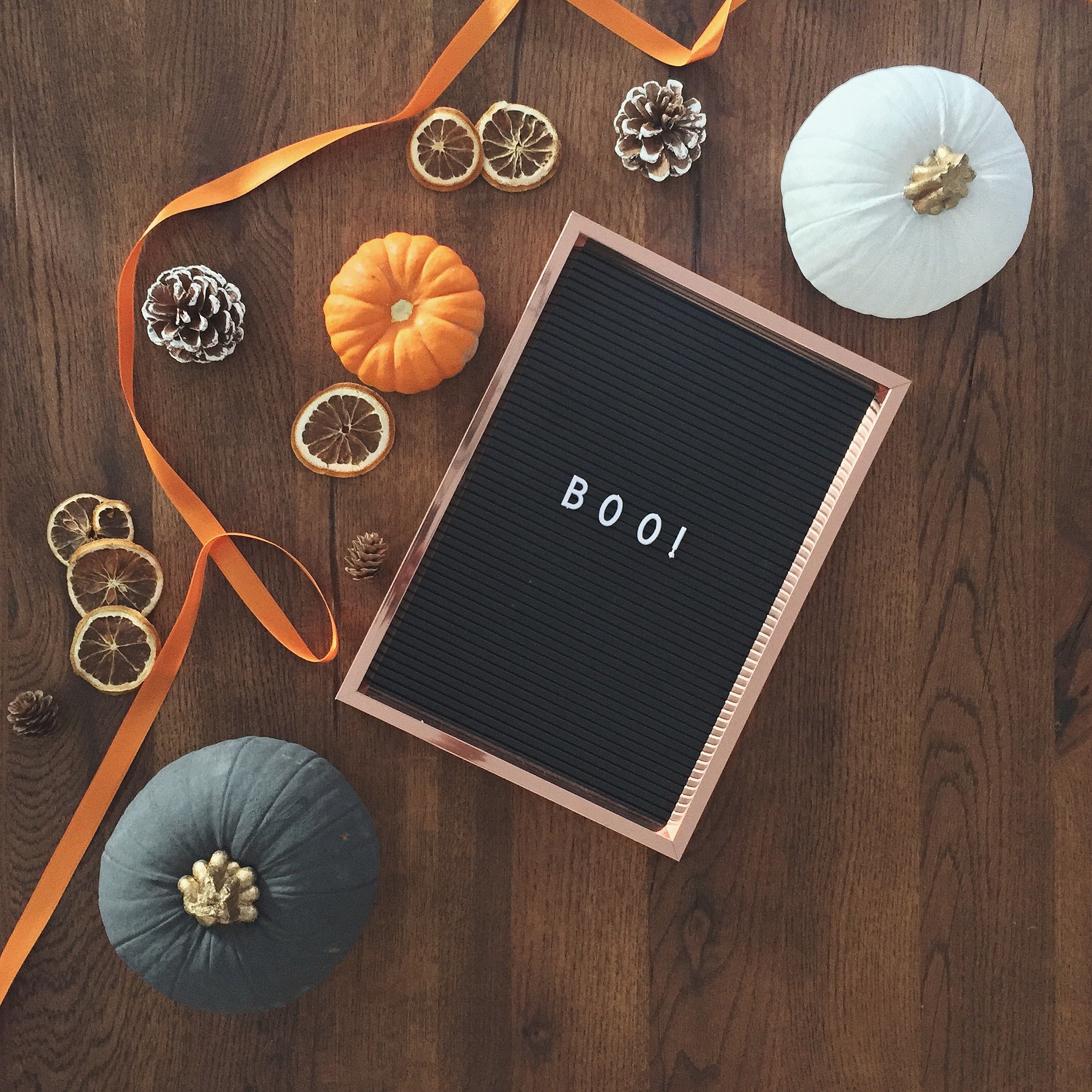 A pegboard reading "boo!" lying flat on a wooden table next to pumpkins of varying colours and sizes.