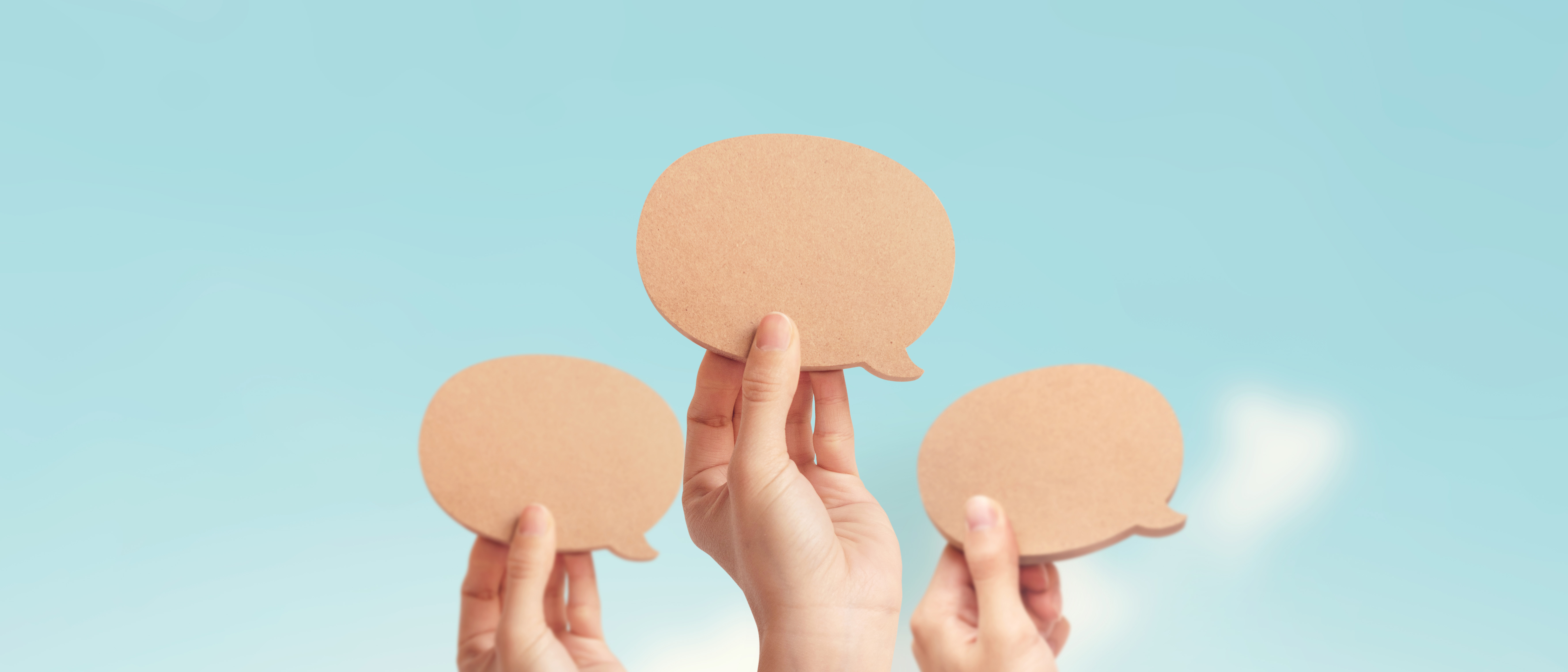 close-up of three hands holding up blank wooden speech bubbles against a pale blue sky
