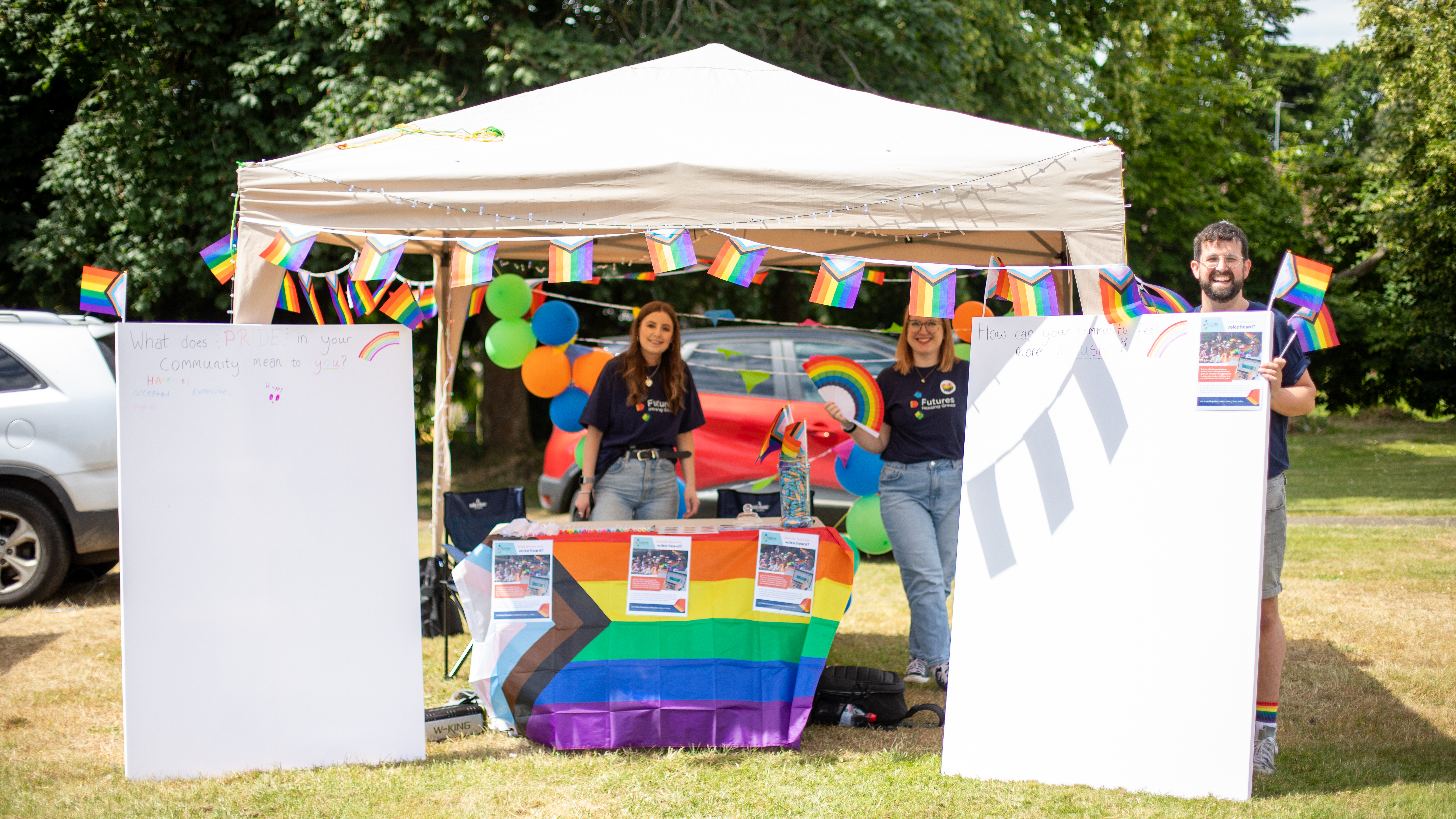 Three members of Futures staff stood around a Pride stall in a Gazebo, flanked with canvases and covered in brightly coloured rainbow flags.