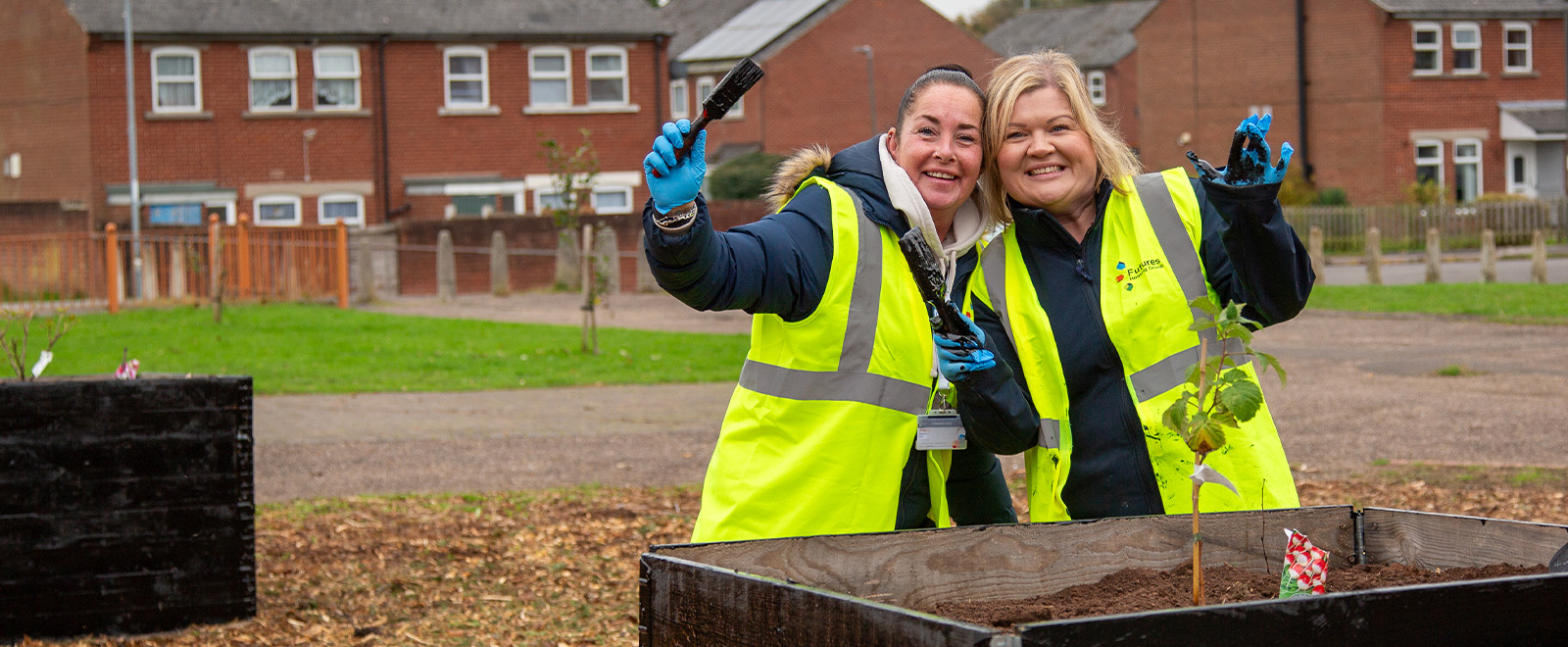 Two smiling people in hi-vis jackets posing for a picture holding paintbrushes in front of a planter