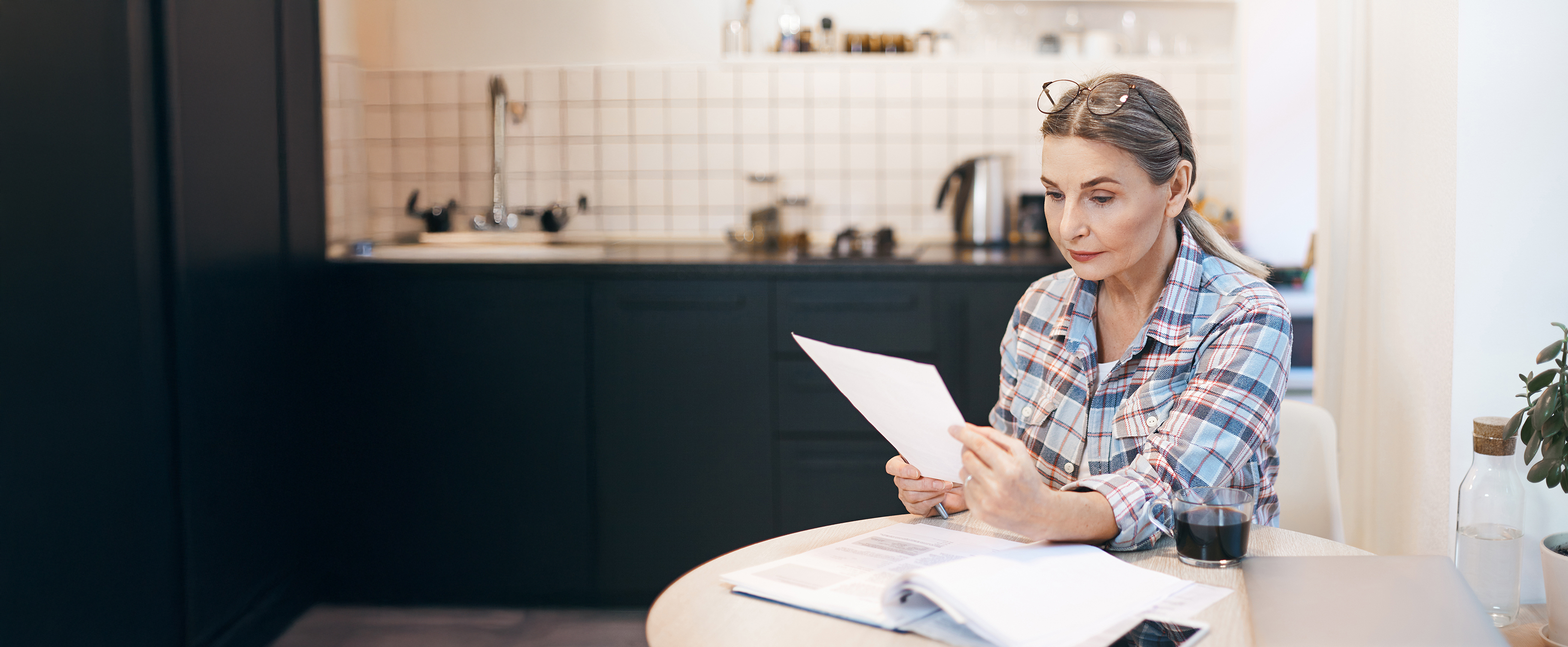 A woman in a plaid shirt sits at a kitchen table, carefully reading a document.